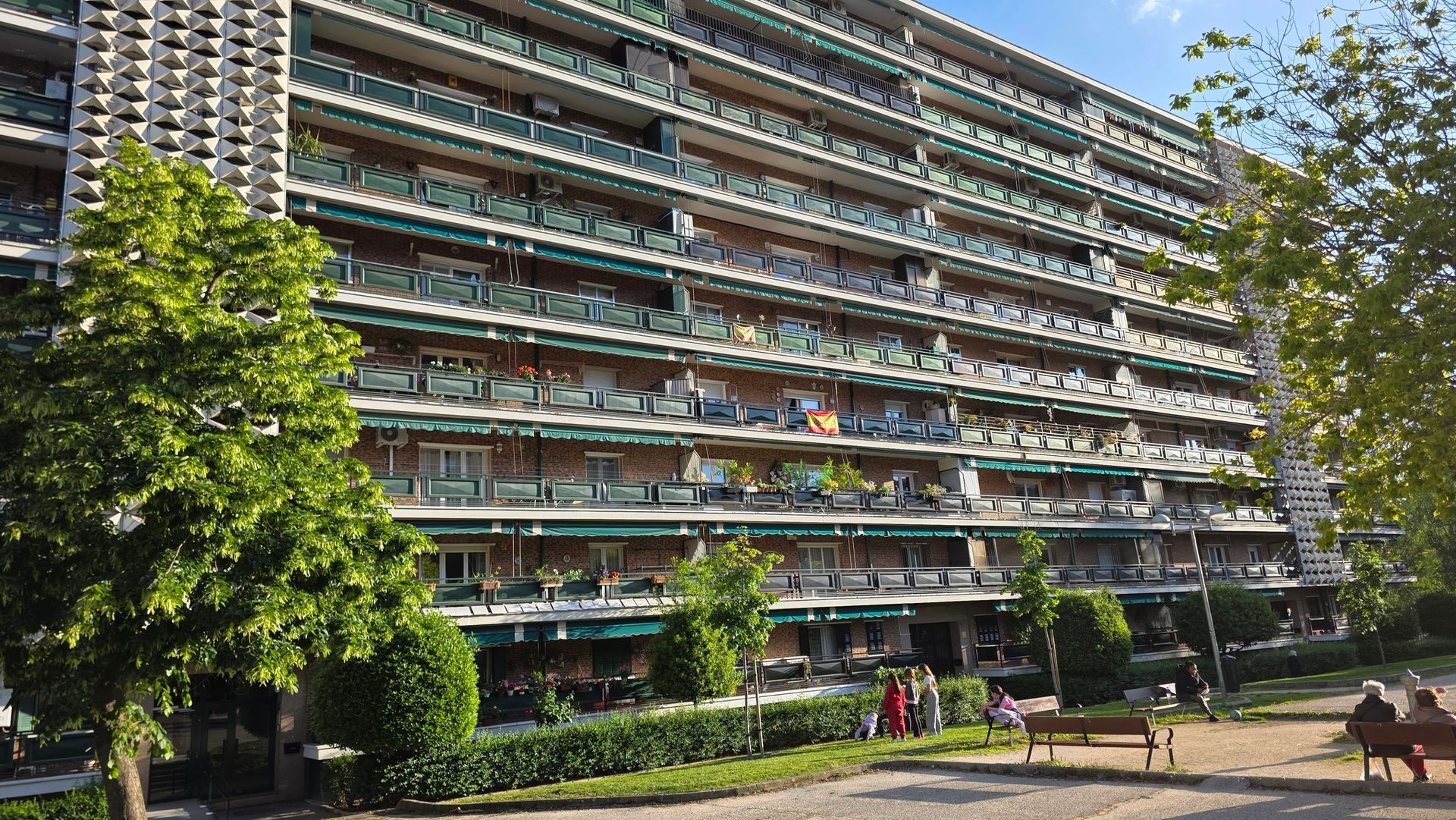 Gran edificio de apartamentos con balcones junto a un sendero arbolado y algunas personas paseando al aire libre.