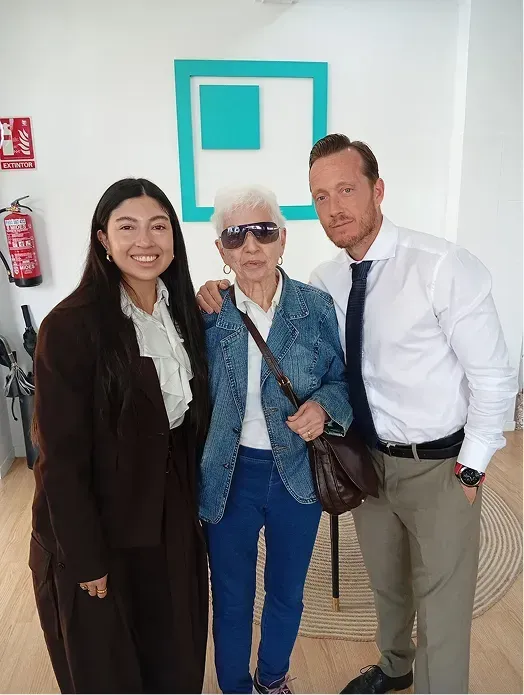 Tres personas posando en el interior, entre ellas una persona mayor con gafas de sol, frente a una pared blanca con un cuadro de color turquesa.