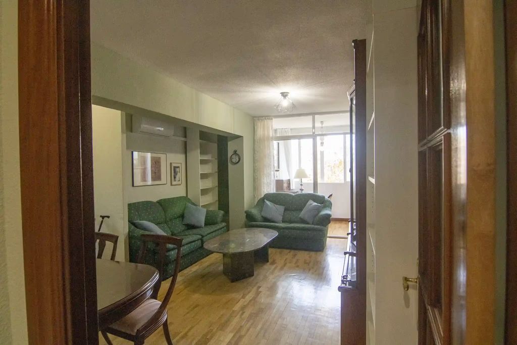 Sala de estar con sofás verdes, suelos de madera y luz natural que entra por un gran ventanal.
