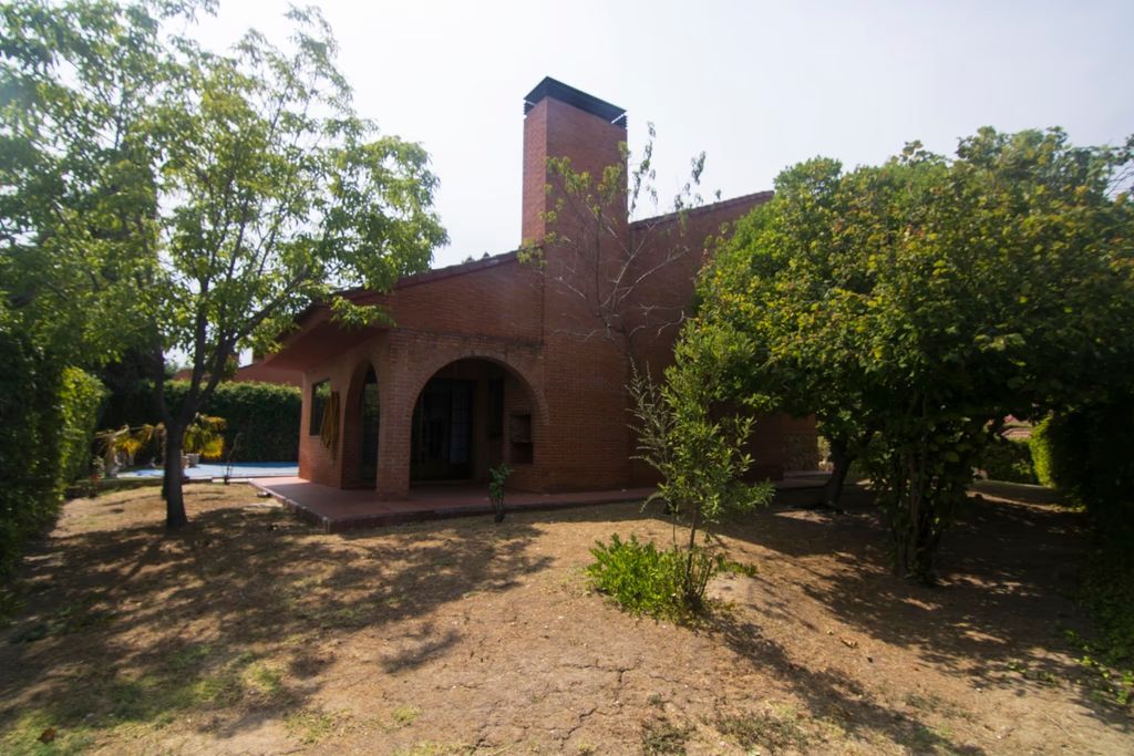 Casa de ladrillo con porche arqueado y chimenea, rodeada de árboles en un patio soleado.