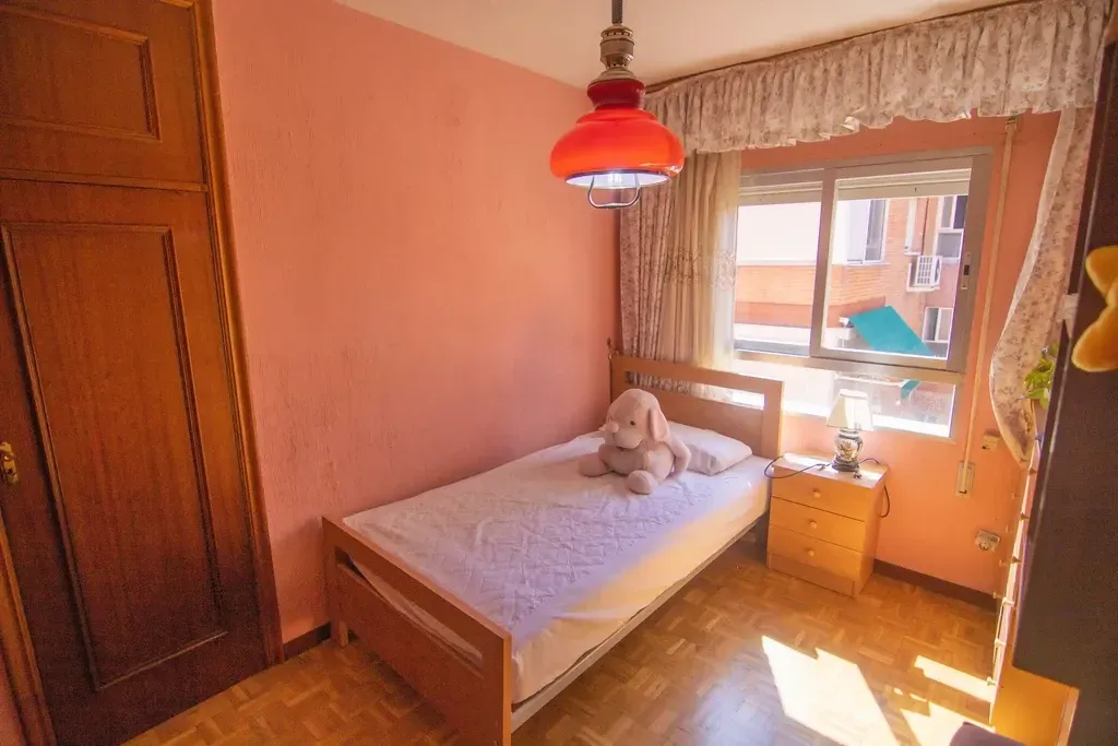 Habitación pequeña con una cama individual, armario de madera, lámpara colgante roja y luz natural que entra por la ventana.