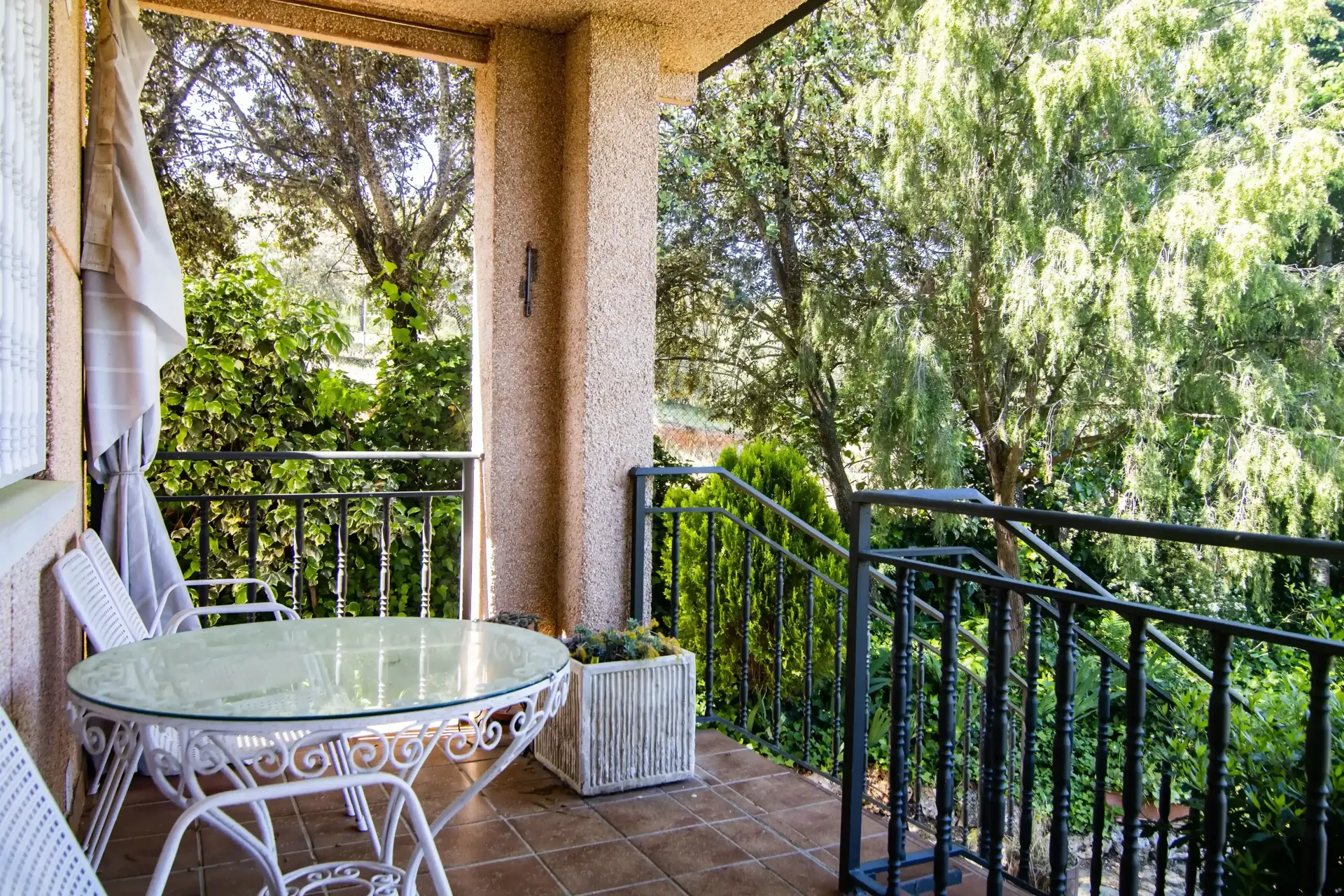 Balcón cubierto con mesa de cristal, sillas blancas y barandilla negra con vistas a frondosos árboles verdes.