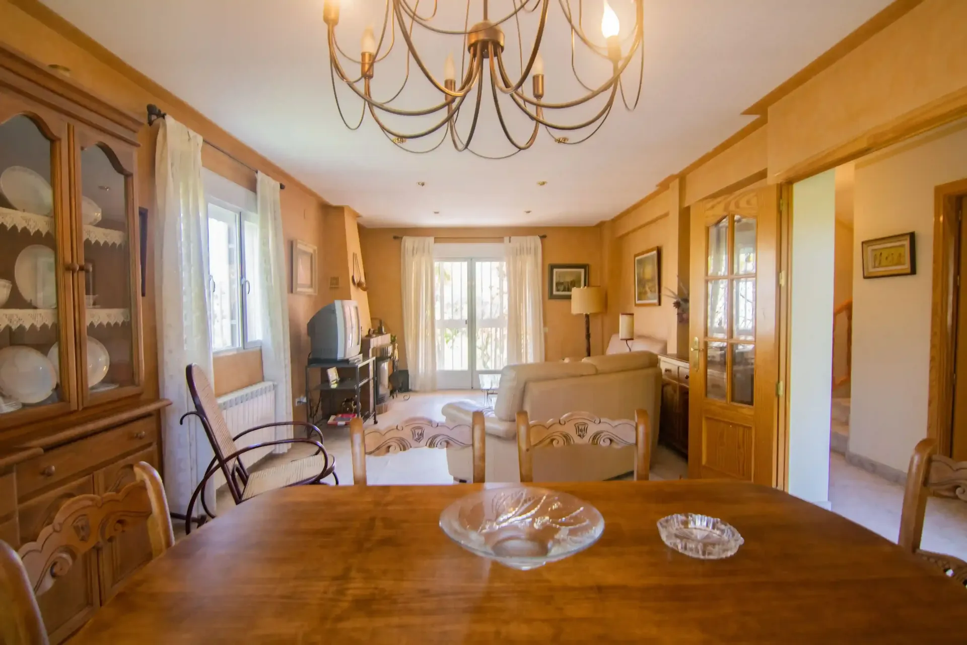 Luminoso salón con paneles de madera, lámpara de araña, chimenea, sofá y grandes ventanales.