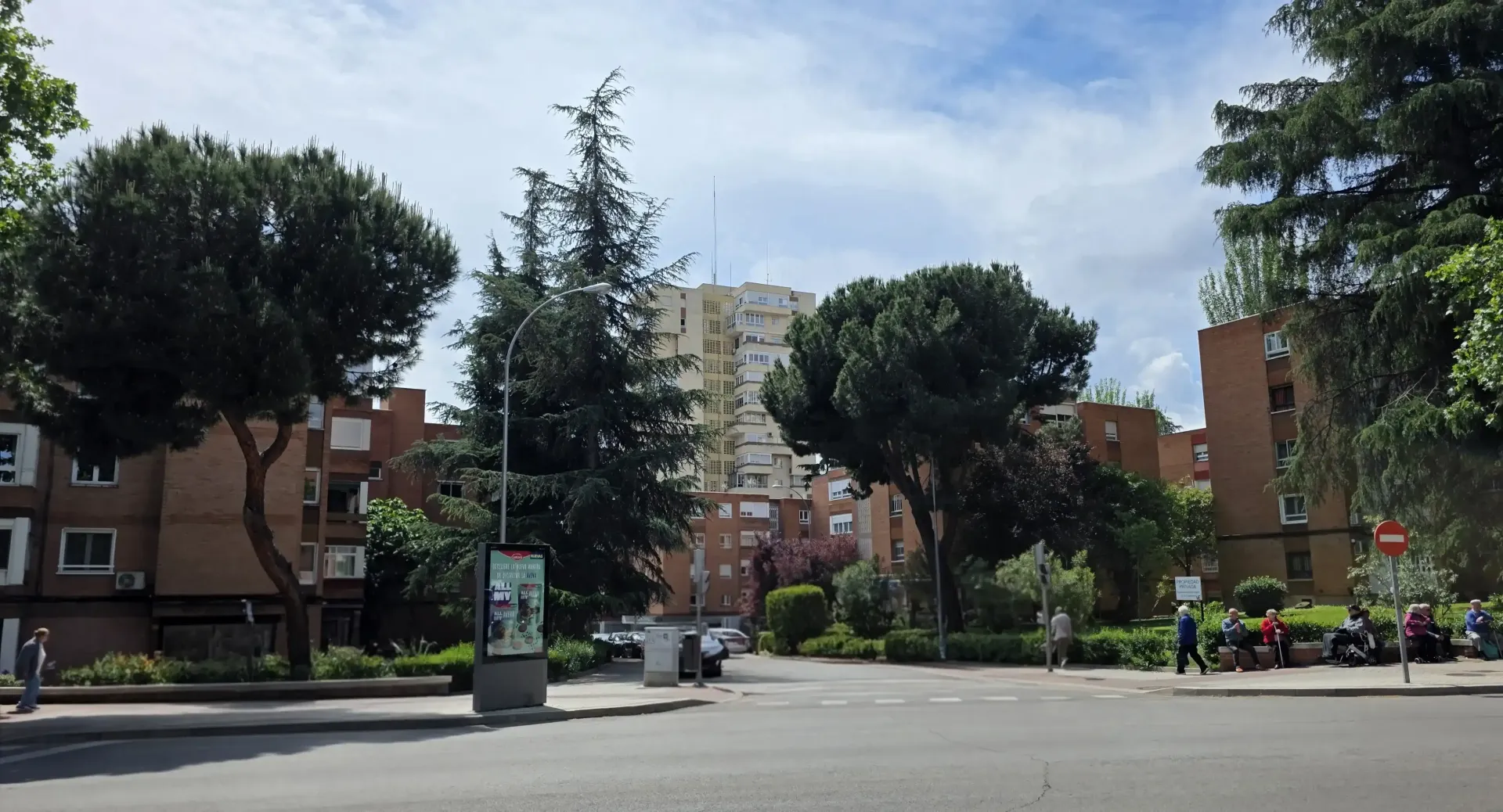Calle urbana con árboles, edificios de apartamentos y gente montando en patinetes en una plaza soleada.