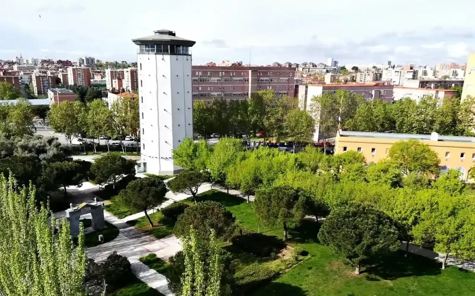 Un parque verde con una alta torre blanca, árboles, senderos y edificios de la ciudad al fondo.