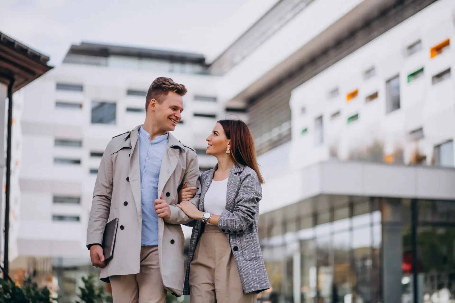 Dos personas caminando y conversando frente a modernos edificios de oficinas, sonriendo y vestidas con abrigos claros.