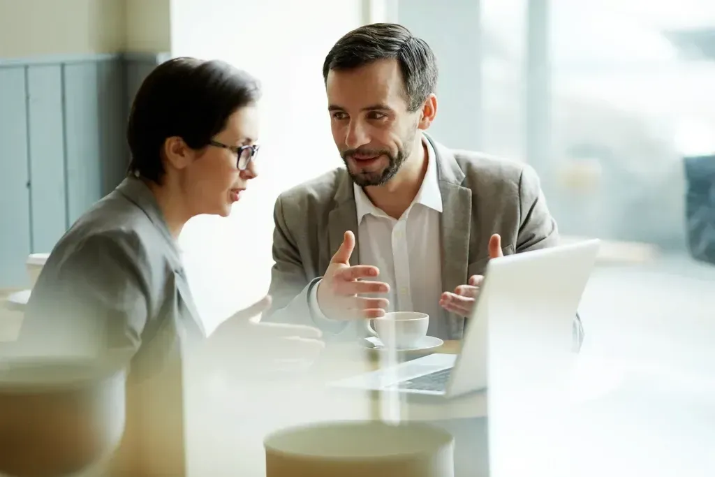Dos compañeros de trabajo conversando en una mesa de una oficina luminosa, con un ordenador portátil abierto cerca.