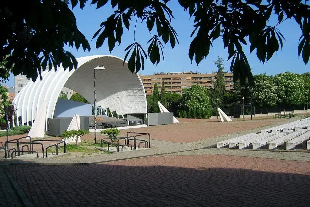 Anfiteatro al aire libre con escenario de concha blanca, filas de asientos y árboles bajo un cielo despejado.