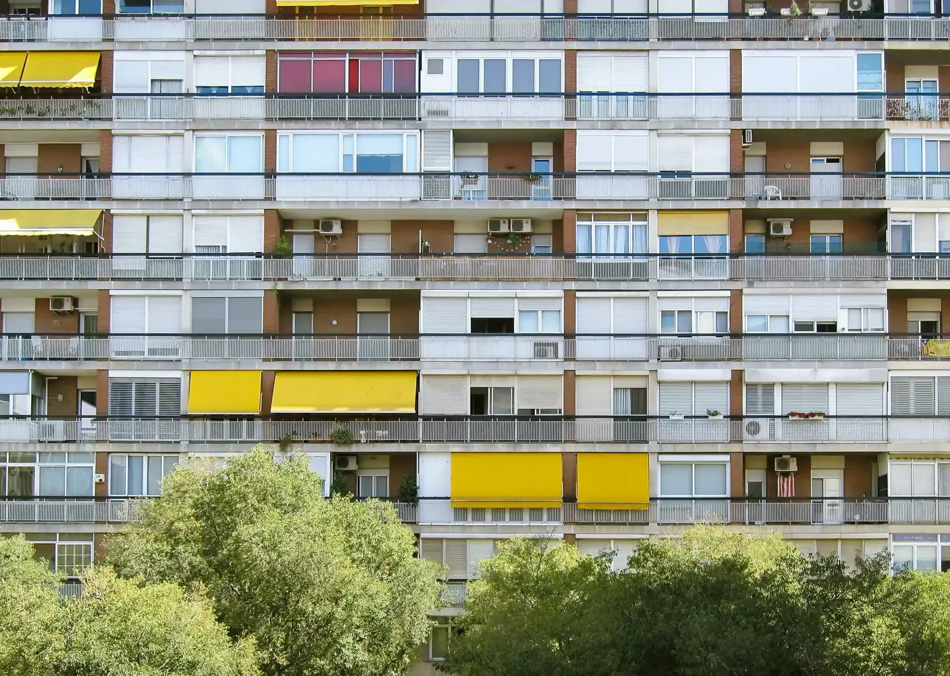Fachada de un edificio de apartamentos con balcones, toldos coloridos y árboles en la fachada.