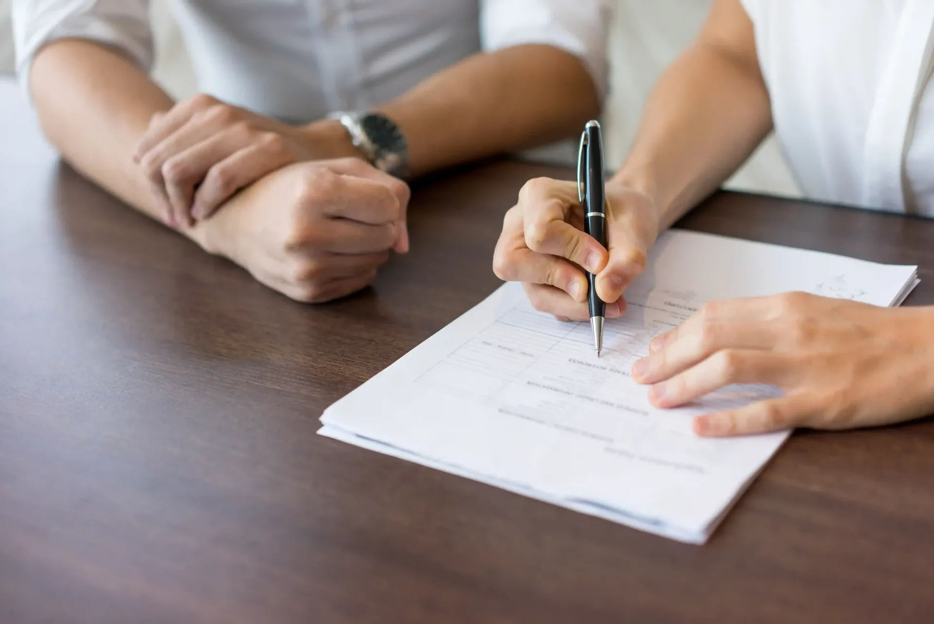 Manos firmando un documento en una mesa de madera con otra persona cerca.