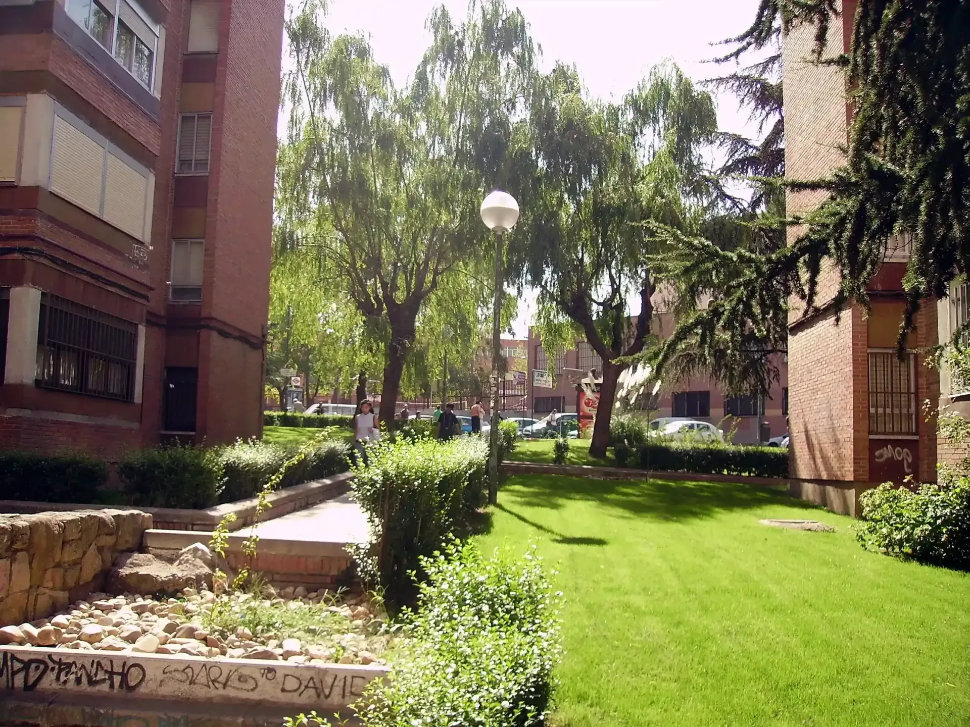 Patio entre edificios de ladrillo con césped verde, árboles y una fuente de piedra en un día soleado.
