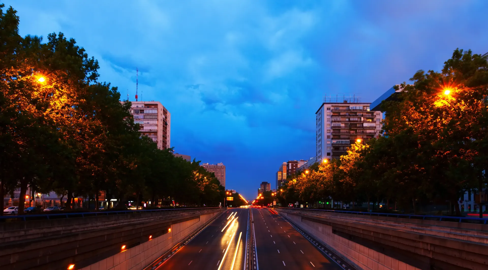 Avenida de la ciudad al atardecer con estelas de luz, árboles y farolas brillantes bajo un cielo azul.