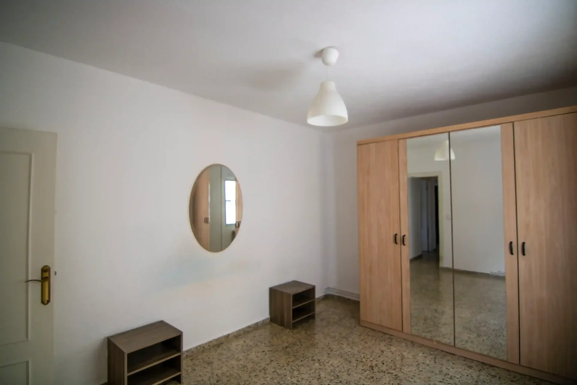 Habitación pequeña con paredes blancas, un armario con espejo, dos mesitas y un espejo de pared ovalado.