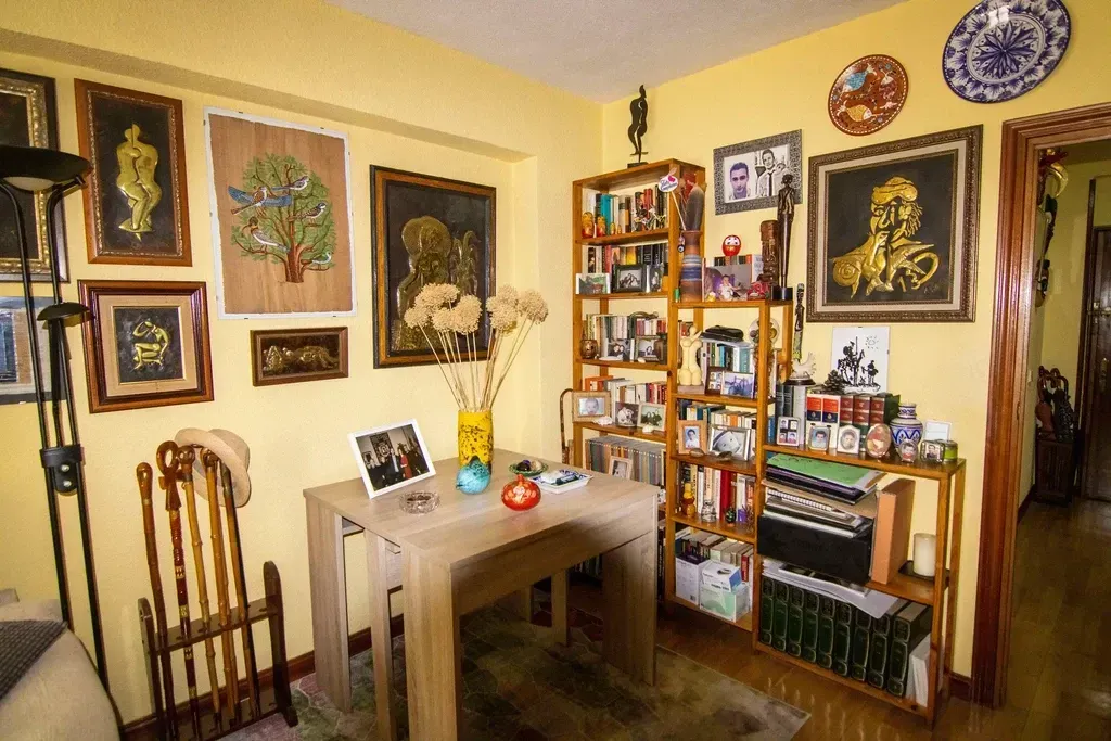 Habitación acogedora con paredes amarillas, cuadros enmarcados, una mesita y estanterías llenas de libros y objetos decorativos.