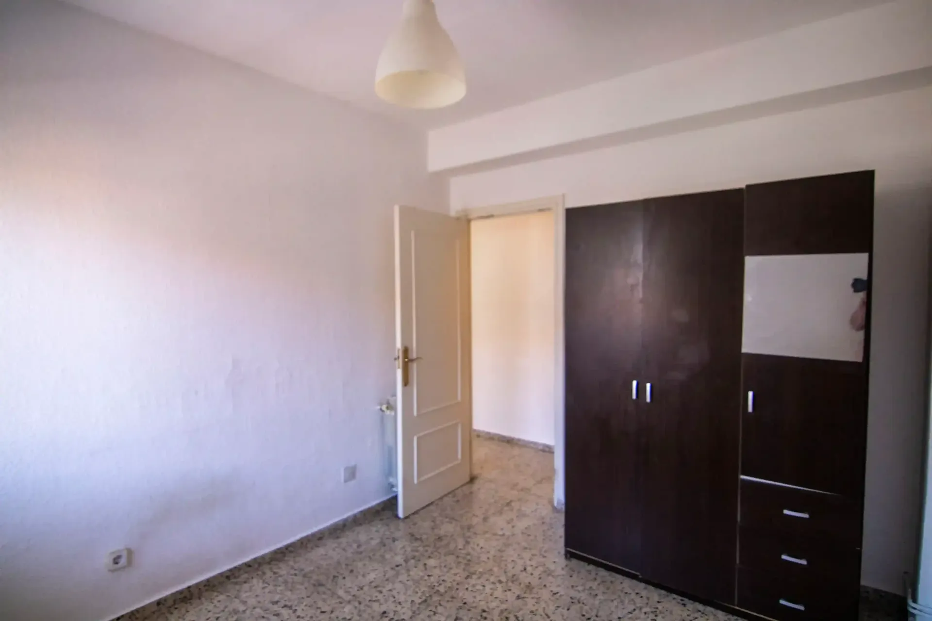 Habitación vacía con paredes blancas, suelo de baldosas, puerta abierta y armario oscuro.