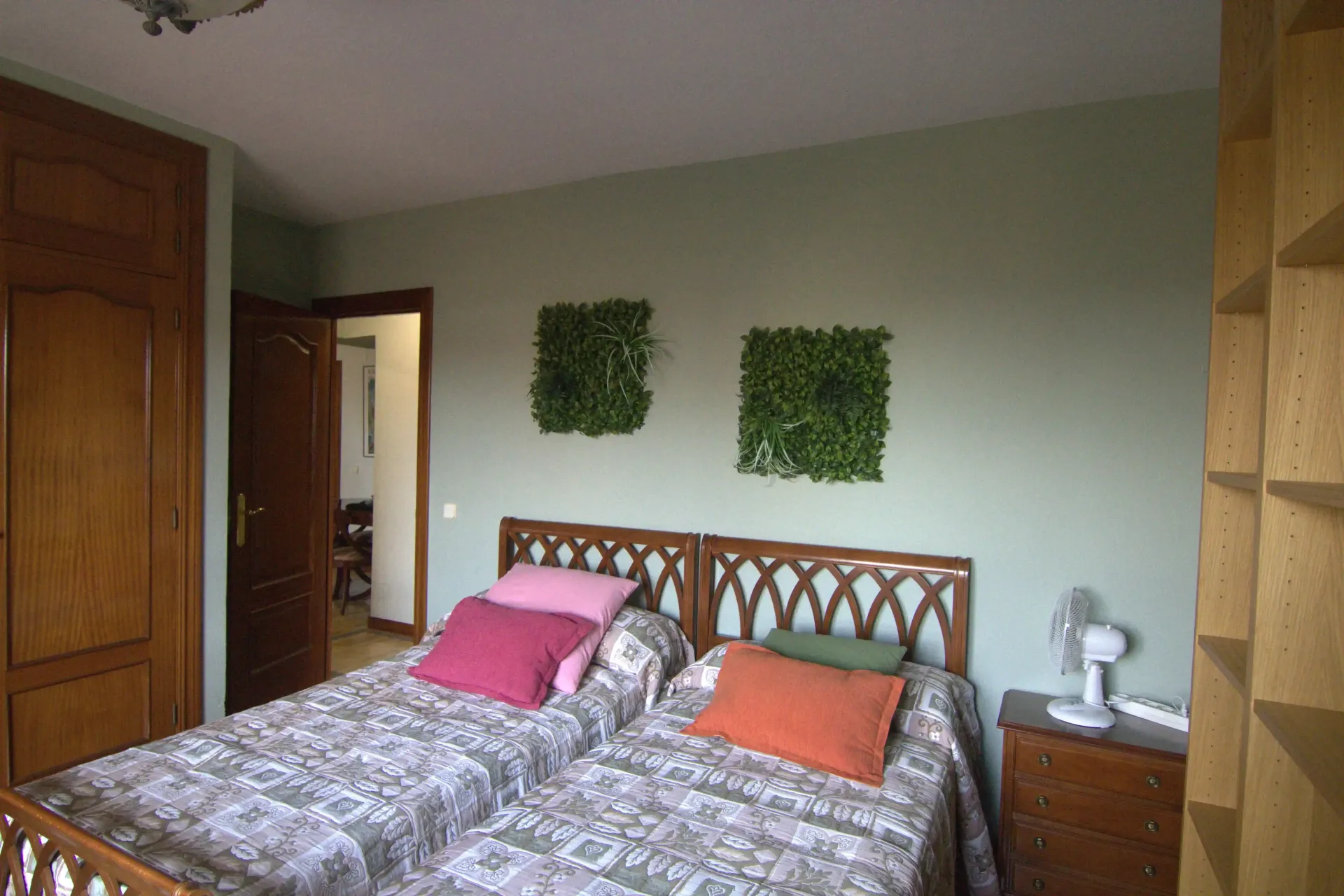 Dormitorio con cama de madera, ropa de cama estampada, almohadas rosas y naranjas y cuadros verdes en la pared.