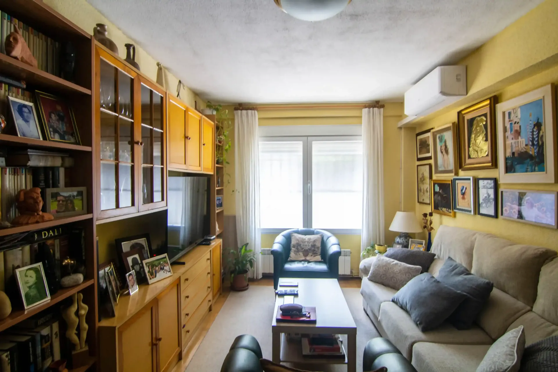 Acogedora sala de estar con sofá gris, armarios de madera, cuadros enmarcados y una luminosa ventana al fondo.