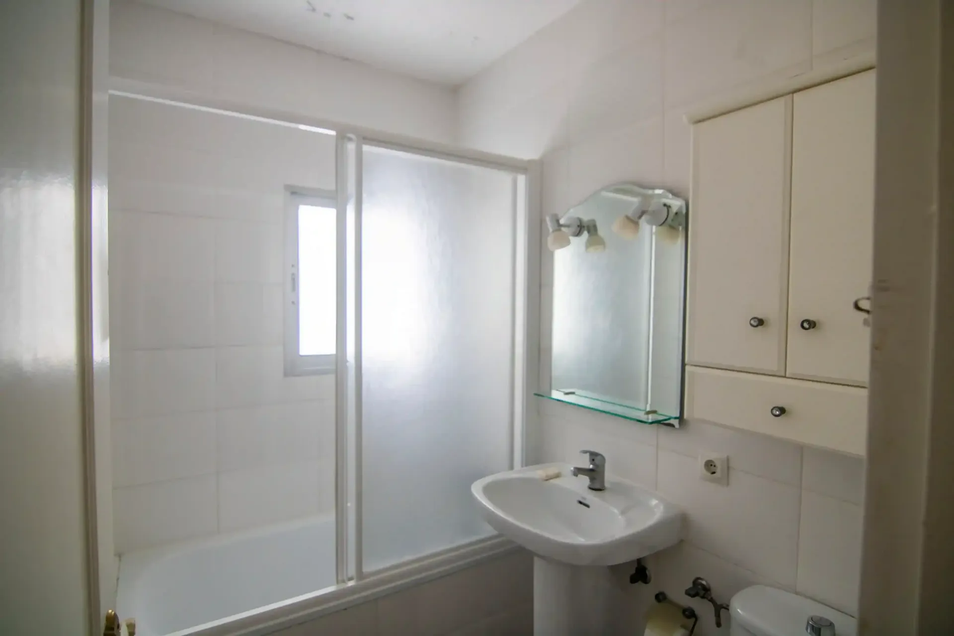 Baño pequeño de color blanco con ducha, lavabo con pedestal, armario con espejo y mampara de ducha esmerilada.
