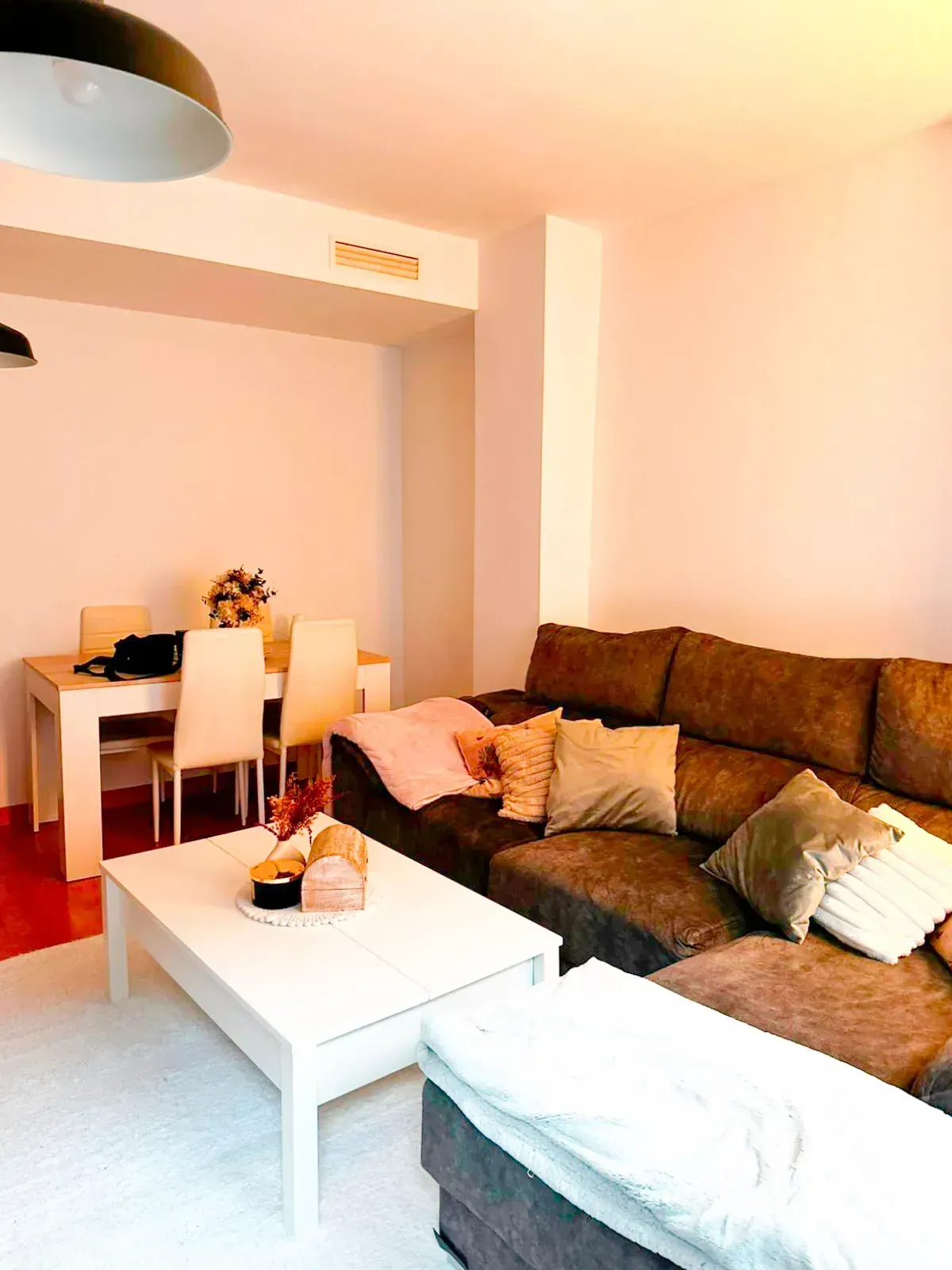Sala de estar luminosa con sofá seccional marrón, mesa de centro blanca y zona de comedor al fondo.