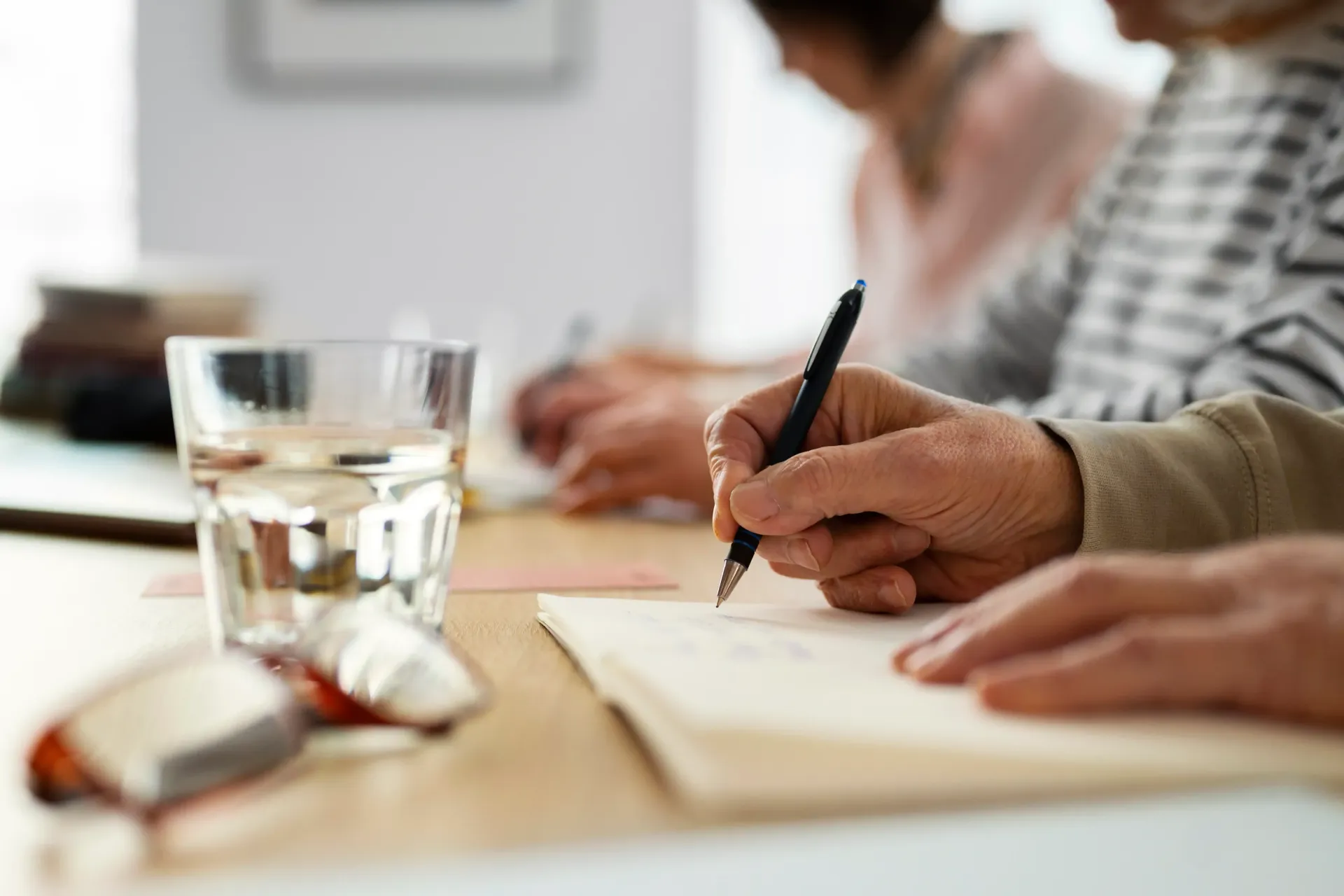 Manos escribiendo en un cuaderno junto a un vaso de agua sobre una mesa, con otras manos borrosas al fondo.