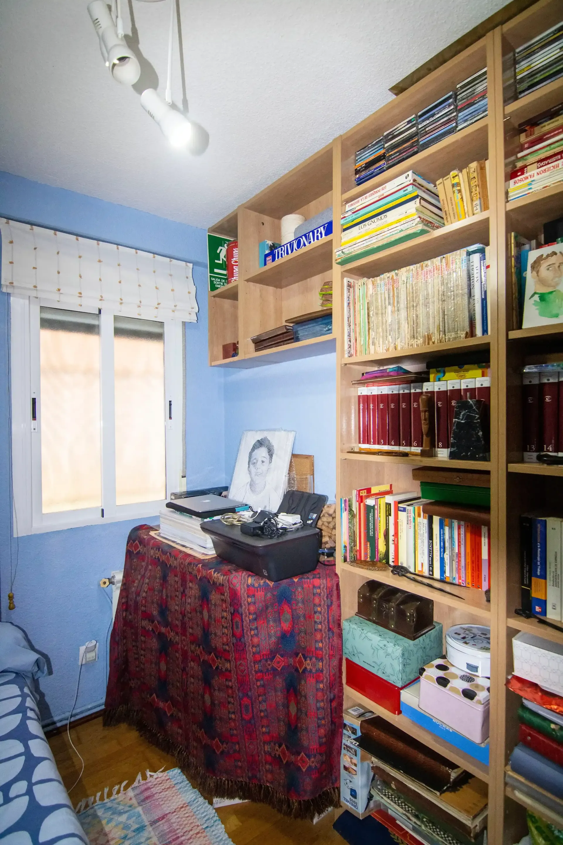Pequeña oficina en casa con estanterías, escritorio, ventana y mesa cubierta con un mantel rojo en una habitación de paredes azules.