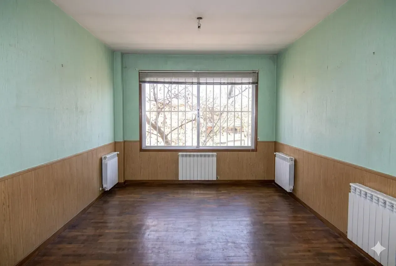 Habitación estrecha y vacía con paredes color menta, suelo de madera, radiador y una gran ventana al fondo.