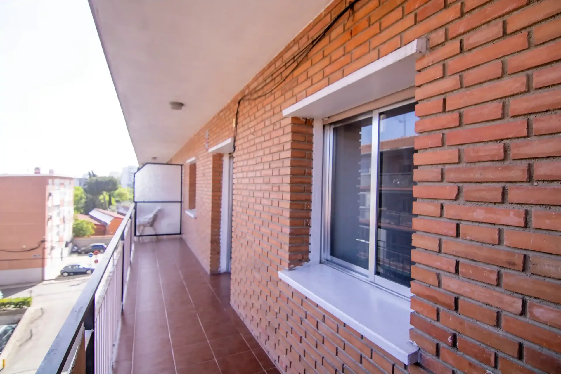 Balcón de apartamento de ladrillo con suelo de baldosas, ventanas correderas y vistas al pasillo.