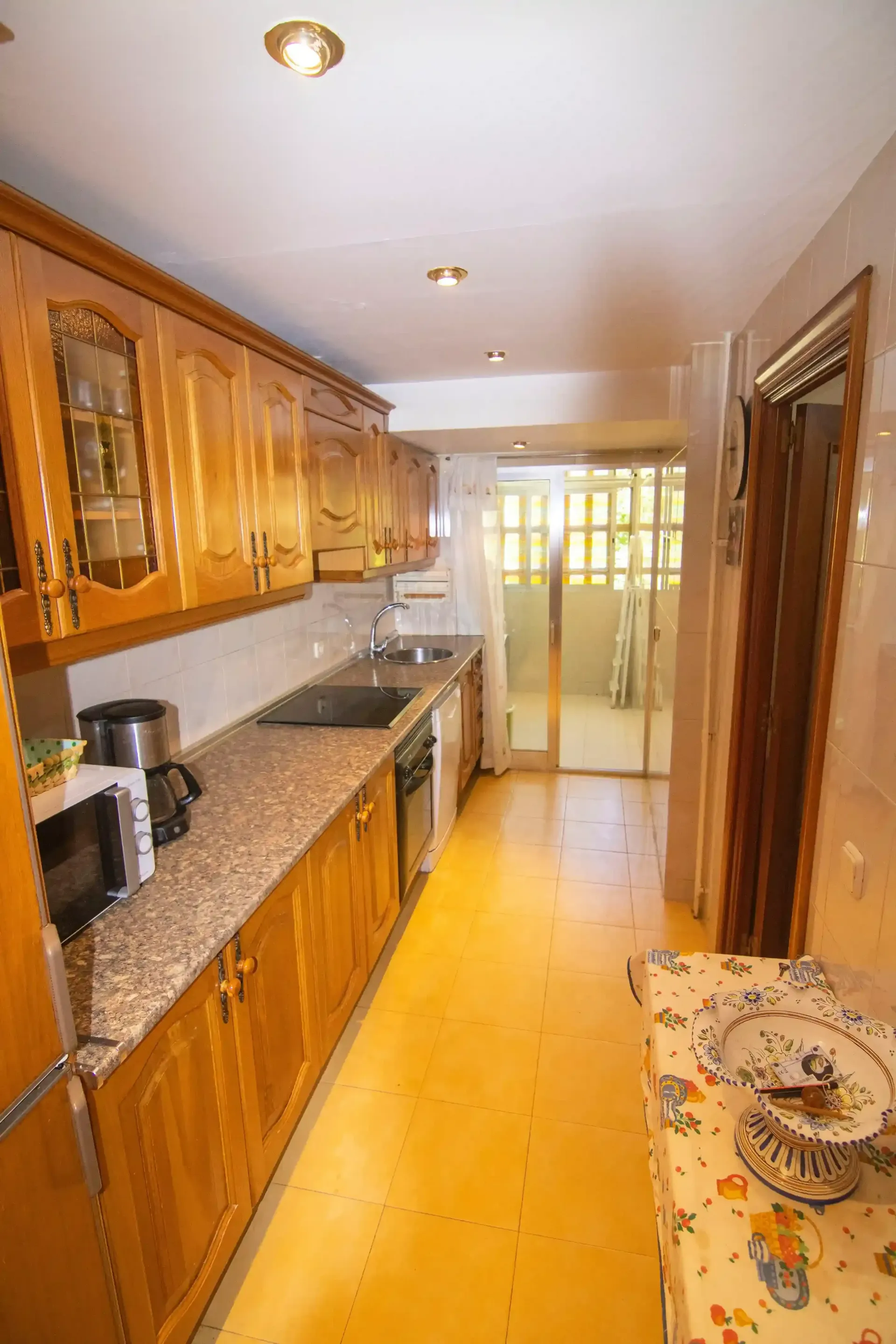 Cocina estrecha con armarios de madera, encimeras de granito y una puerta luminosa al fondo.