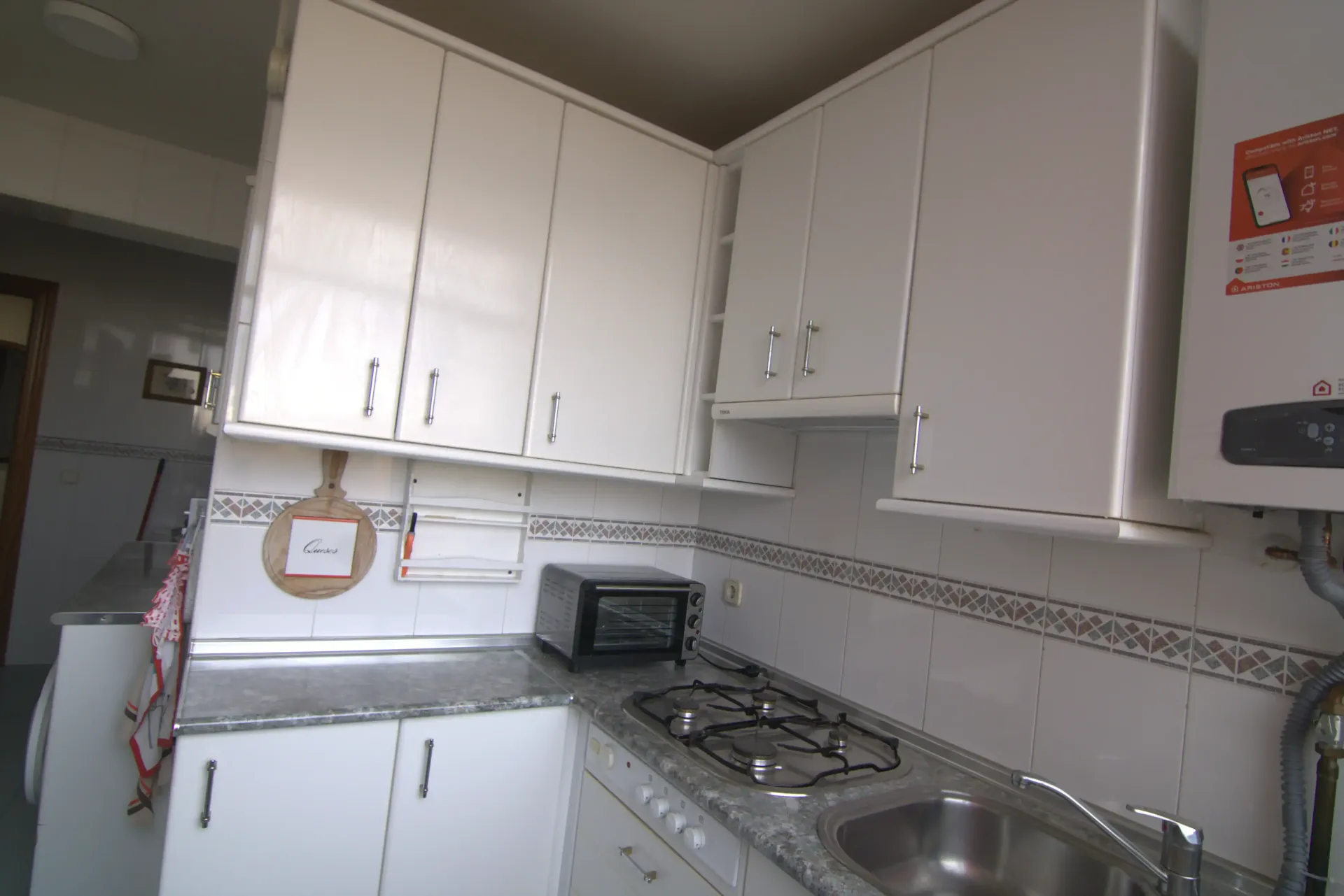 Pequeña cocina blanca con armarios, estufa de gas, fregadero y salpicadero de azulejos.
