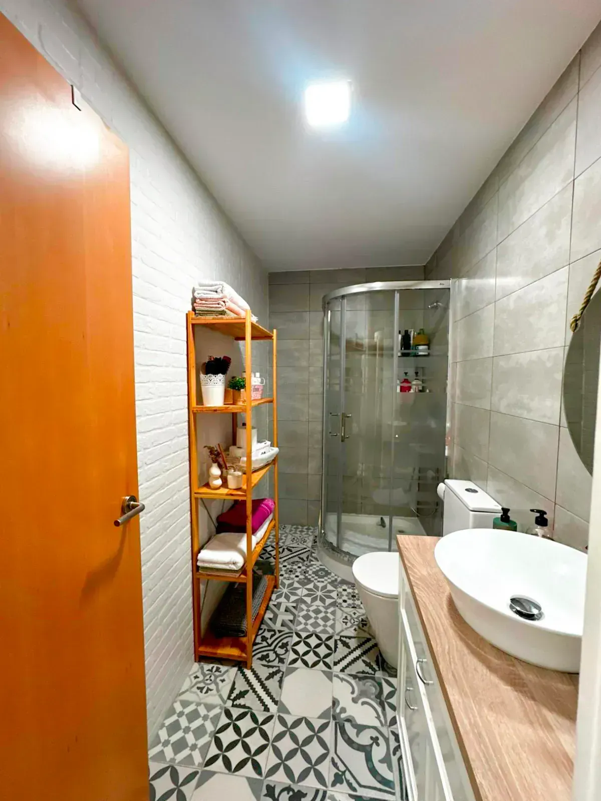 Baño moderno y estrecho con ducha alicatada, estante de madera, lavabo y baldosas geométricas en el suelo.