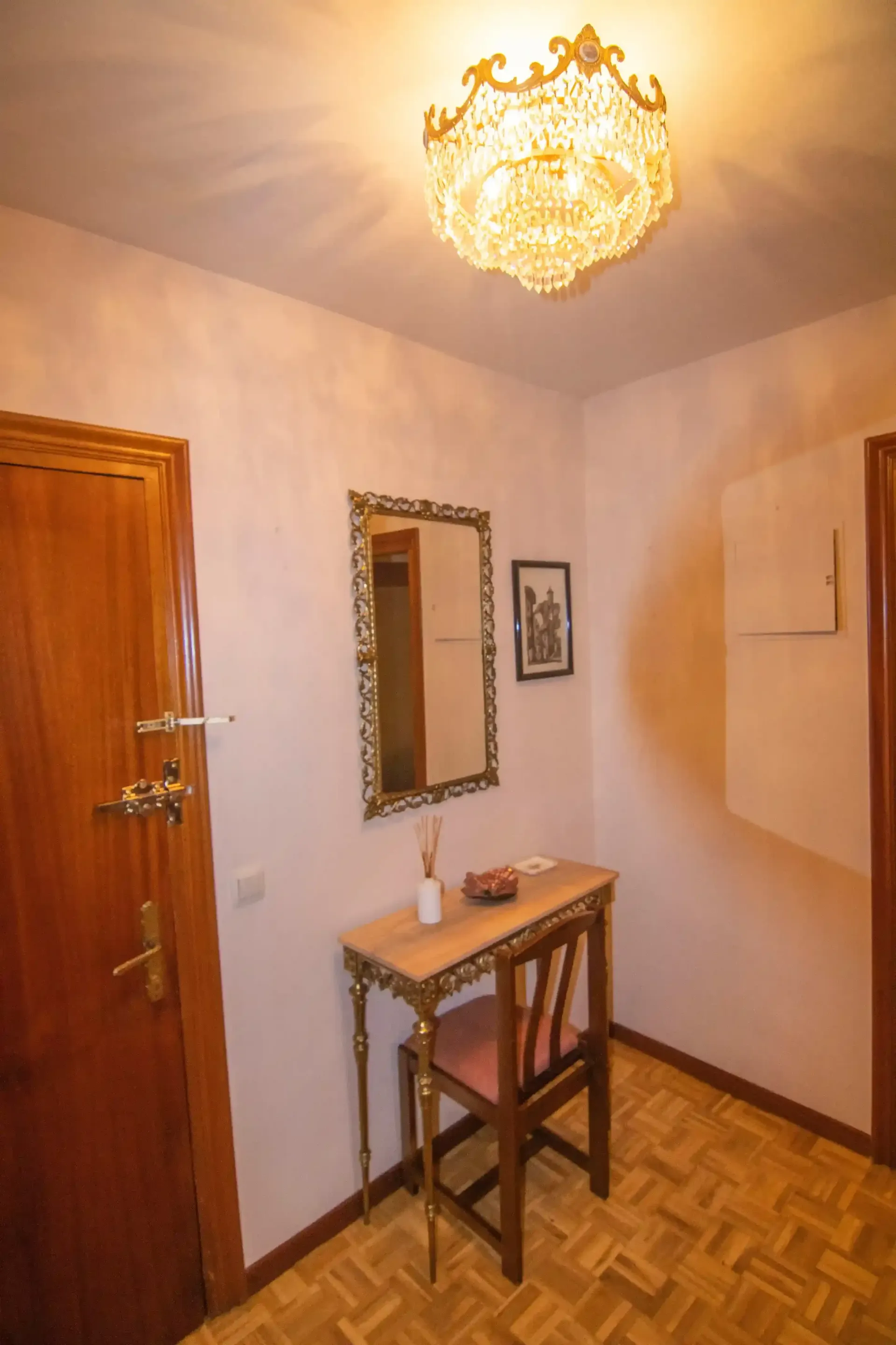 Pequeño pasillo con lámpara de araña, puerta de madera, espejo de pared y consola estrecha sobre suelo de parqué.
