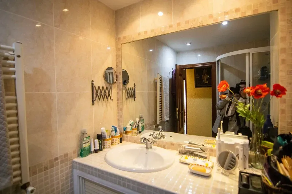 Mueble de baño con espejo grande, azulejos beige, lavabo blanco, artículos de aseo y flores rojas sobre la encimera.