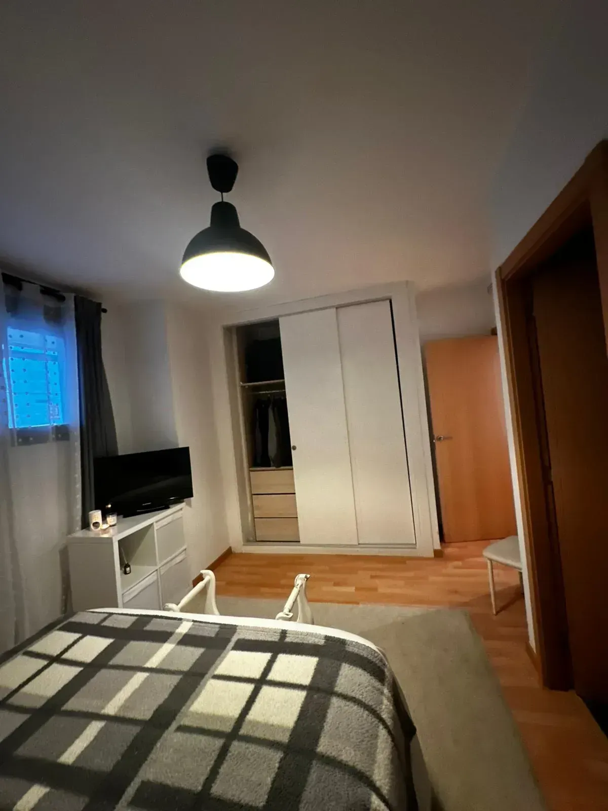 Habitación pequeña con cama, armario, televisor con soporte y lámpara de techo colgante.