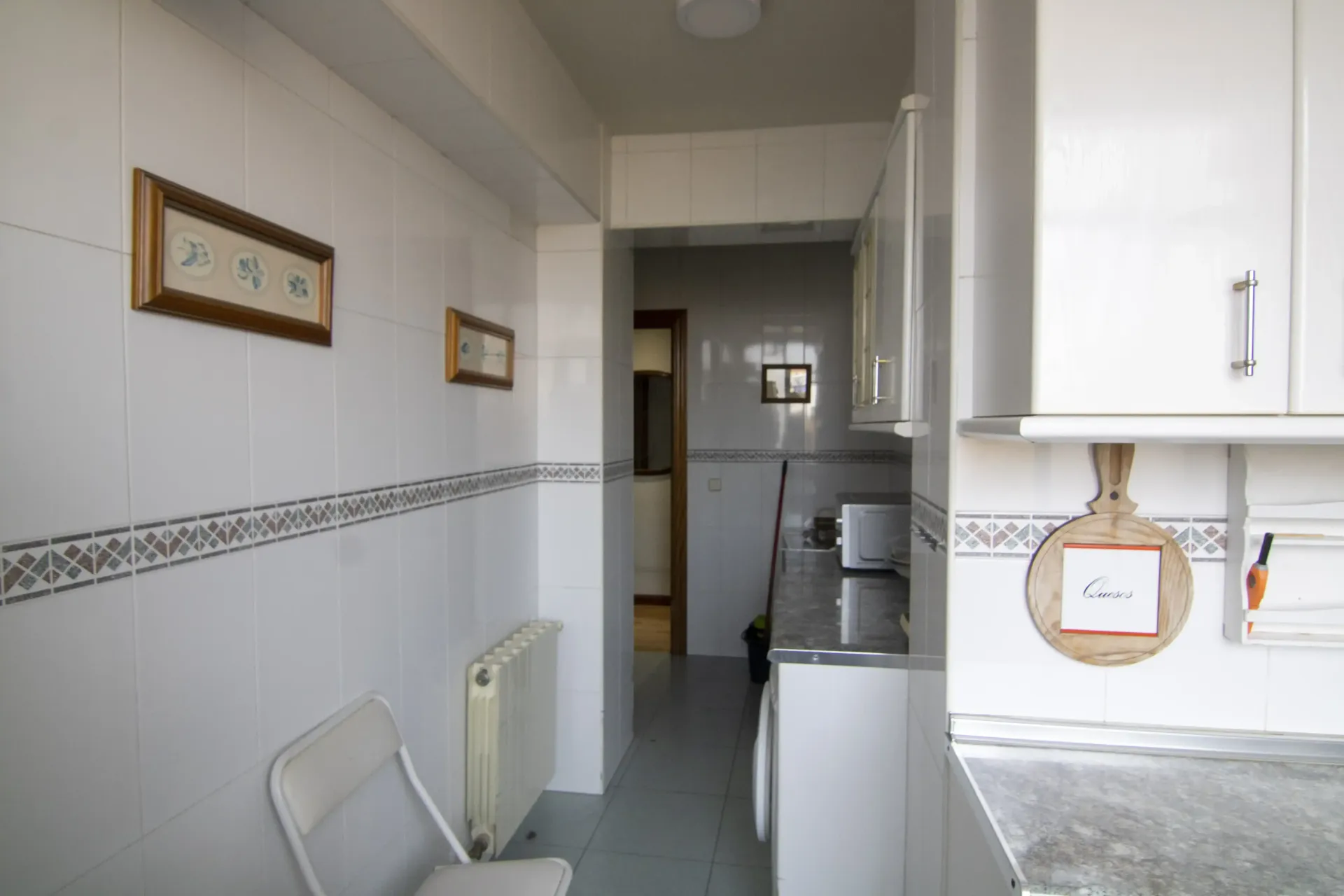 Cocina estrecha de color blanco con paredes alicatadas, fregadero, radiador y armarios a lo largo de un pequeño pasillo.