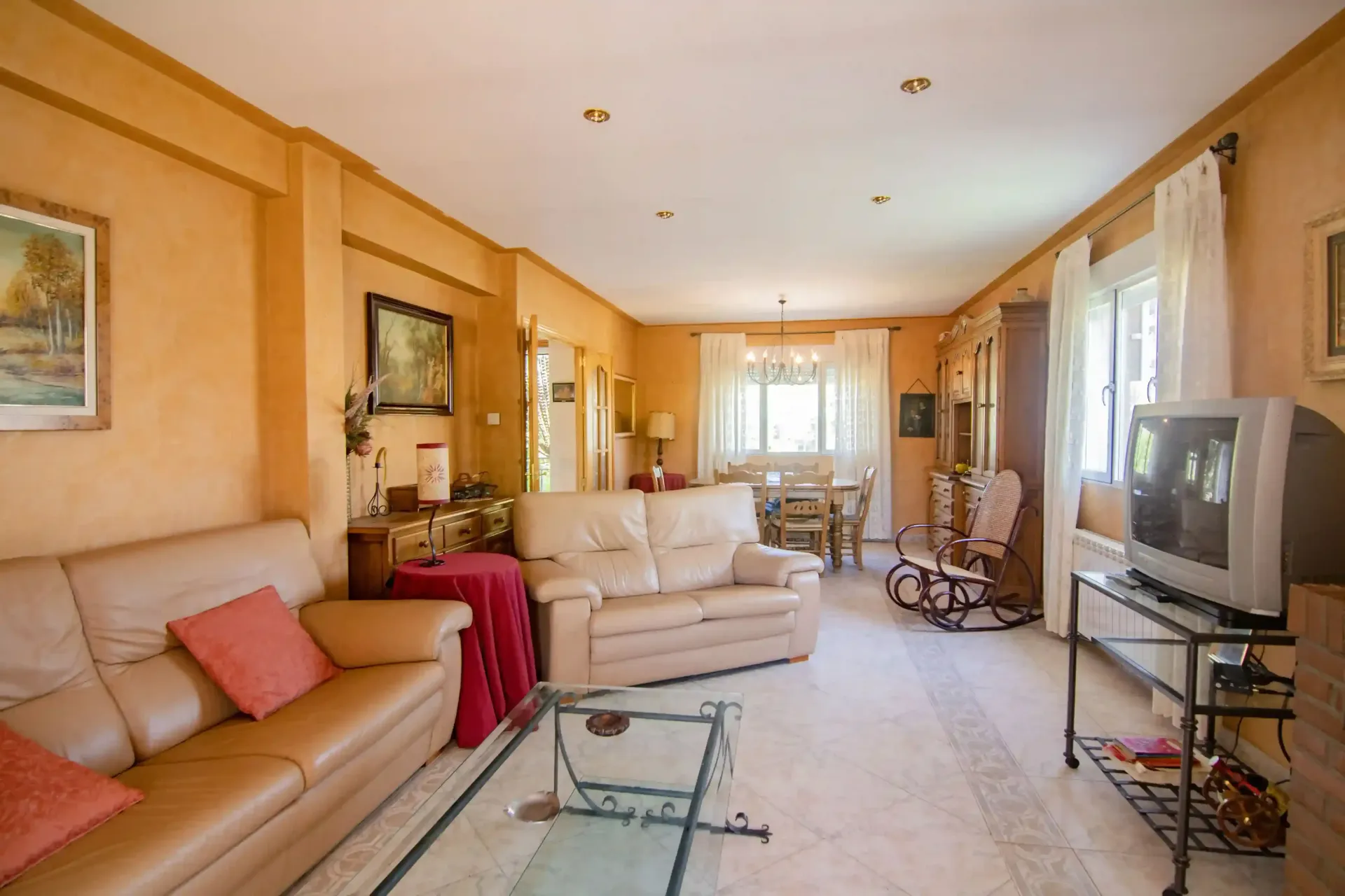 Luminoso salón con sofás beige, televisor, cuadros enmarcados y luz natural que entra por los grandes ventanales.