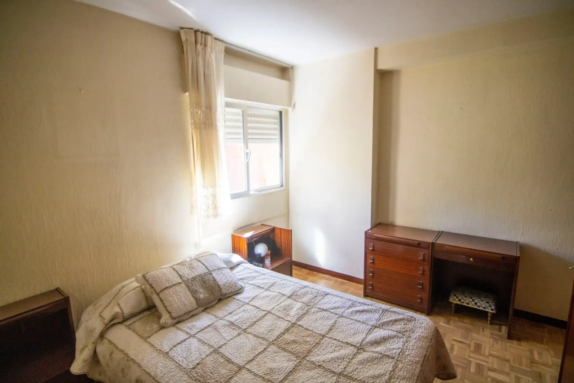 Habitación pequeña con cama sin hacer, ventana, escritorio y cómoda de madera a lo largo de paredes de color beige.