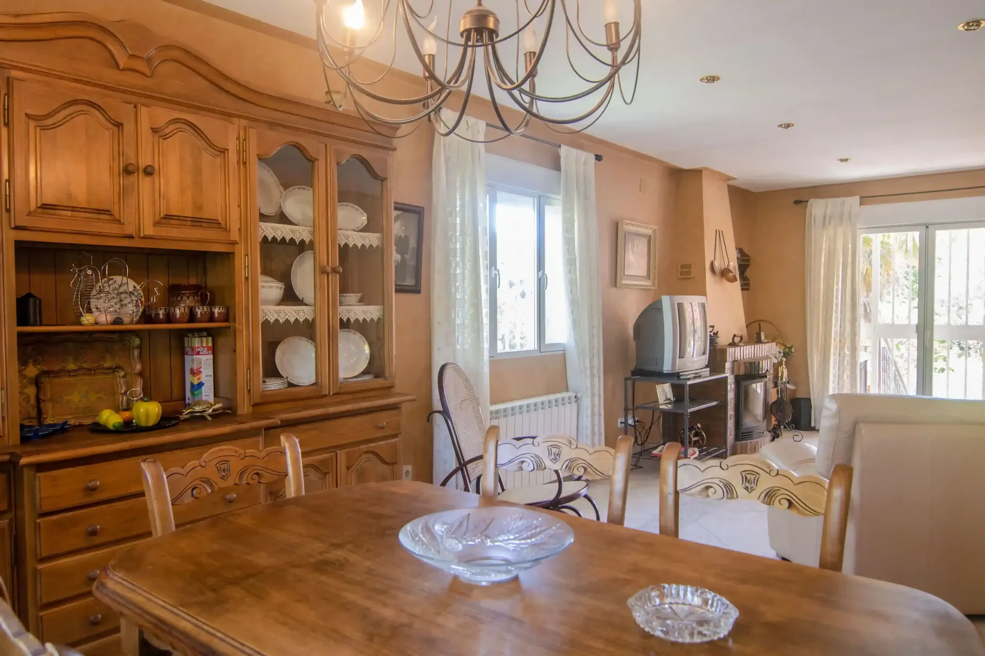 Comedor con paneles de madera, vitrina, lámpara de araña y una gran mesa de madera cerca de ventanas luminosas.