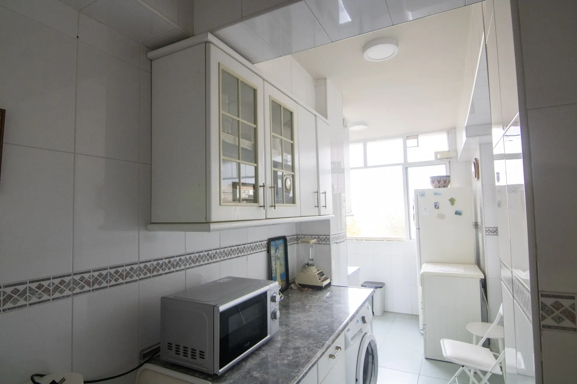 Cocina blanca y luminosa con microondas, encimera, armarios y frigorífico cerca de una puerta con ventana.