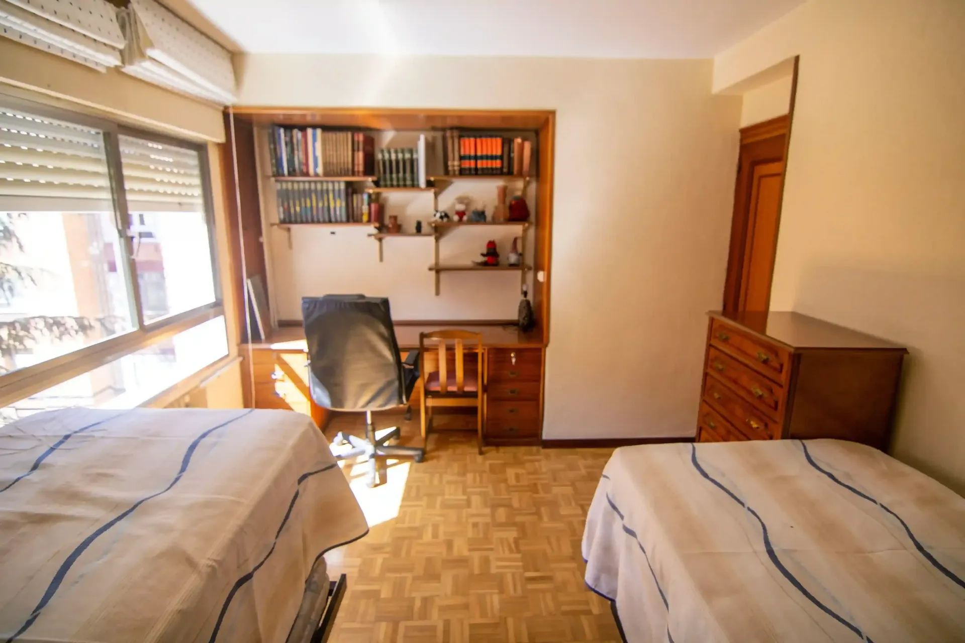 Habitación pequeña con dos camas, un escritorio, estanterías y una ventana con persianas.