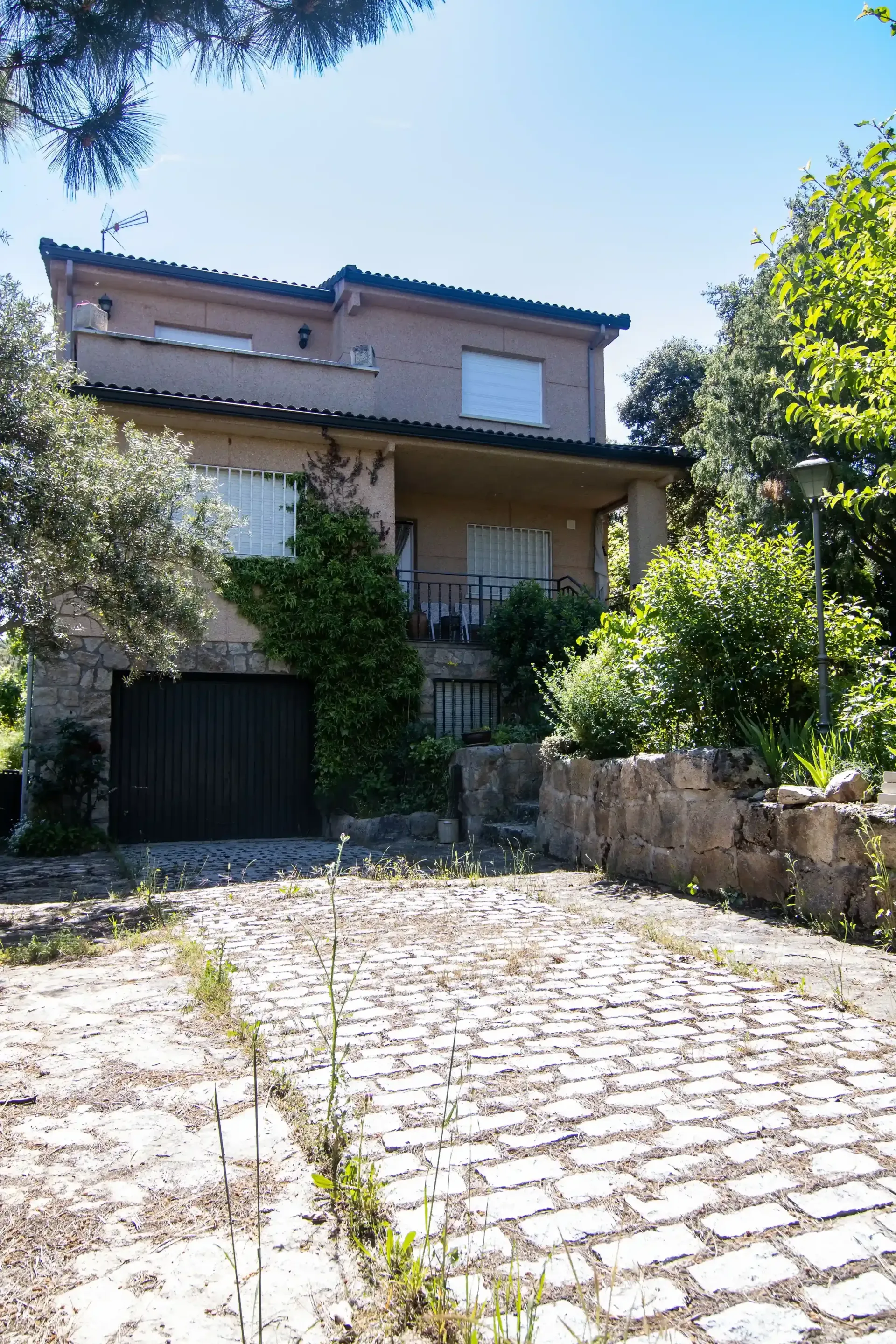Patio de piedra que conduce a una casa de dos pisos con hiedra, un balcón y exuberante vegetación bajo un cielo azul.