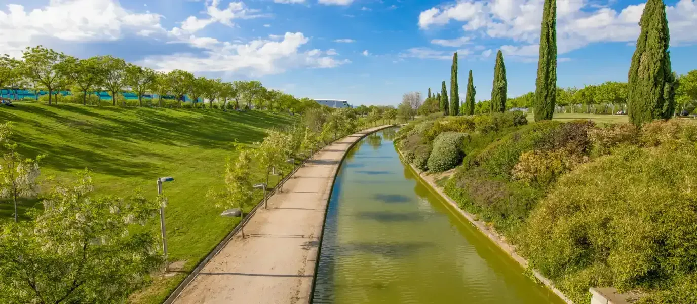 Un estrecho canal junto a un sendero que atraviesa una ladera verde bajo un cielo azul con nubes.
