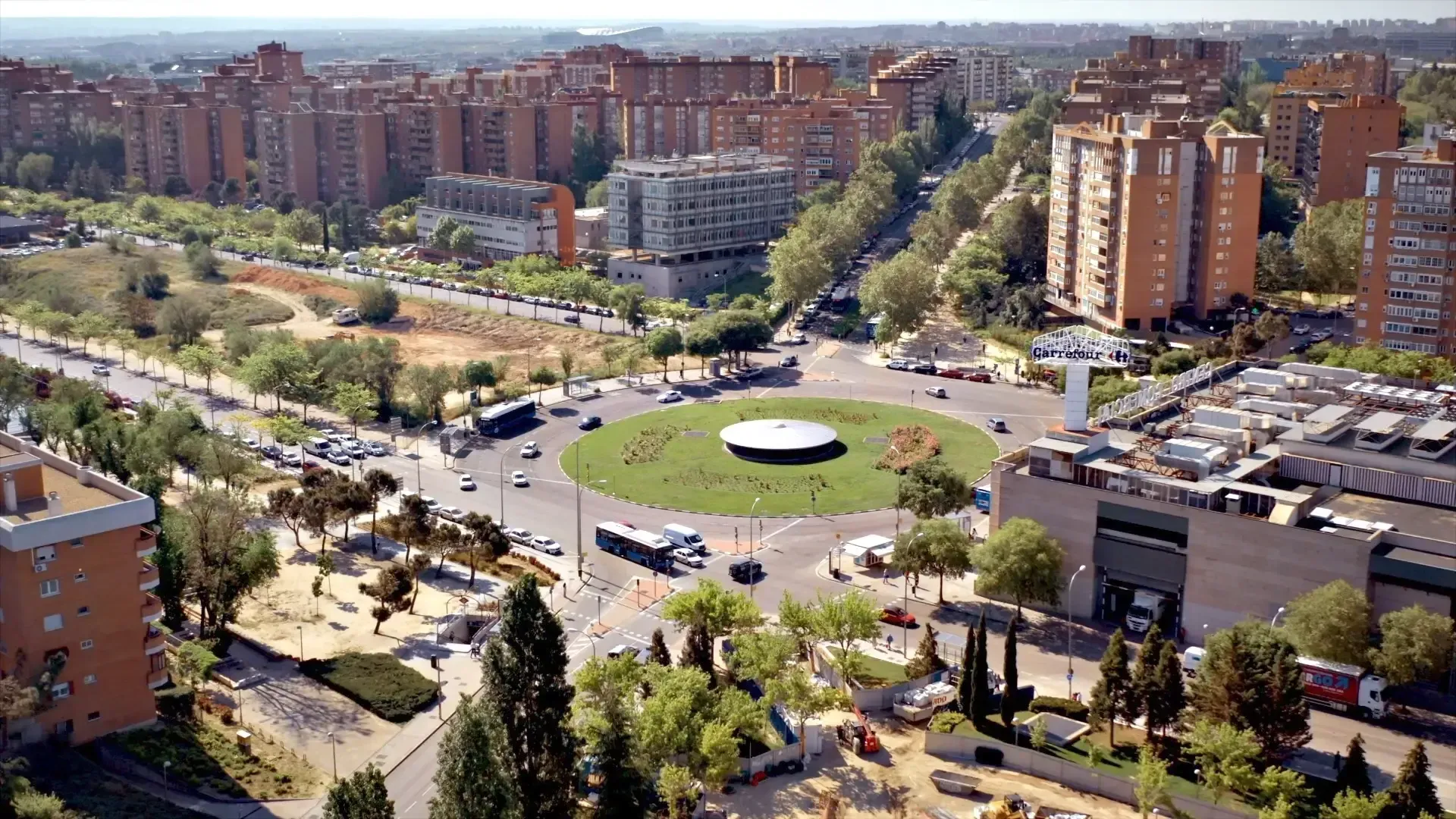 Vista aérea de una rotonda urbana con un centro cubierto de césped, rodeada de carreteras, edificios y árboles.