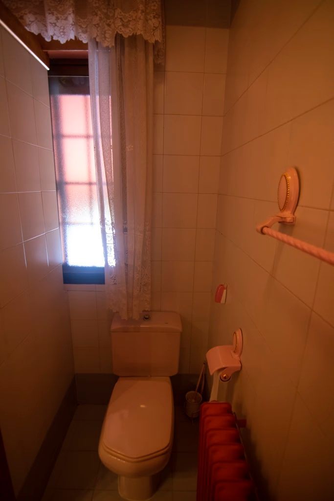 Baño estrecho con inodoro, ventana, paredes alicatadas y un toallero con toallas rojas.