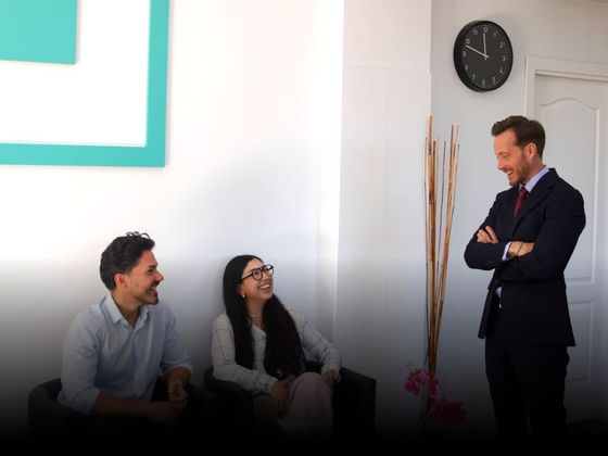 Tres compañeros de trabajo conversando en una oficina moderna; uno de ellos está de pie con los brazos cruzados junto a un reloj de pared.
