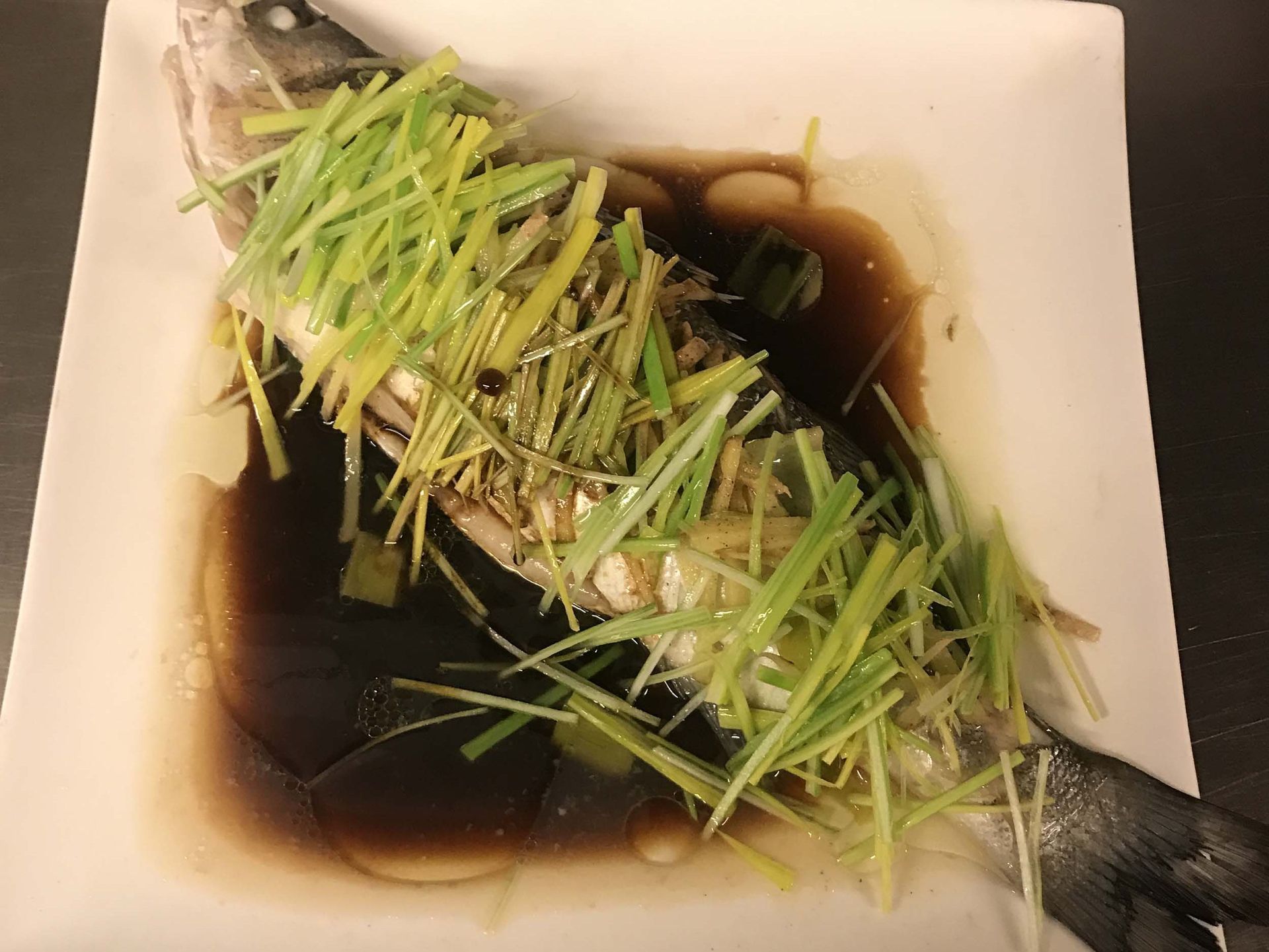 Ein Fisch ist mit Frühlingszwiebeln und Soße bedeckt und liegt auf einem weißen Teller.