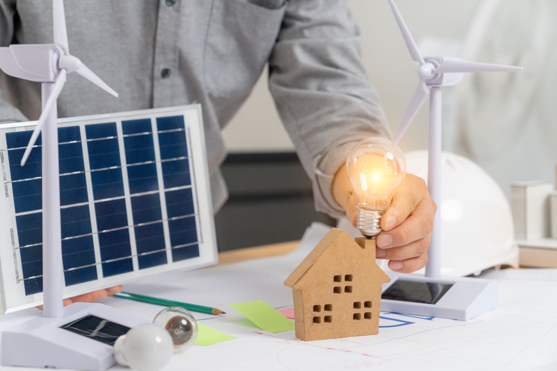 Personne tenant une ampoule allumée à côté d'une maquette de maison, d'une éolienne miniature et d'un panneau solaire sur un bureau.
