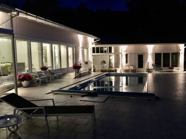 Yönäkymä valkoisesta talosta ja uima-altaasta, joka on valaistu. Patiolla on lepotuoleja.
