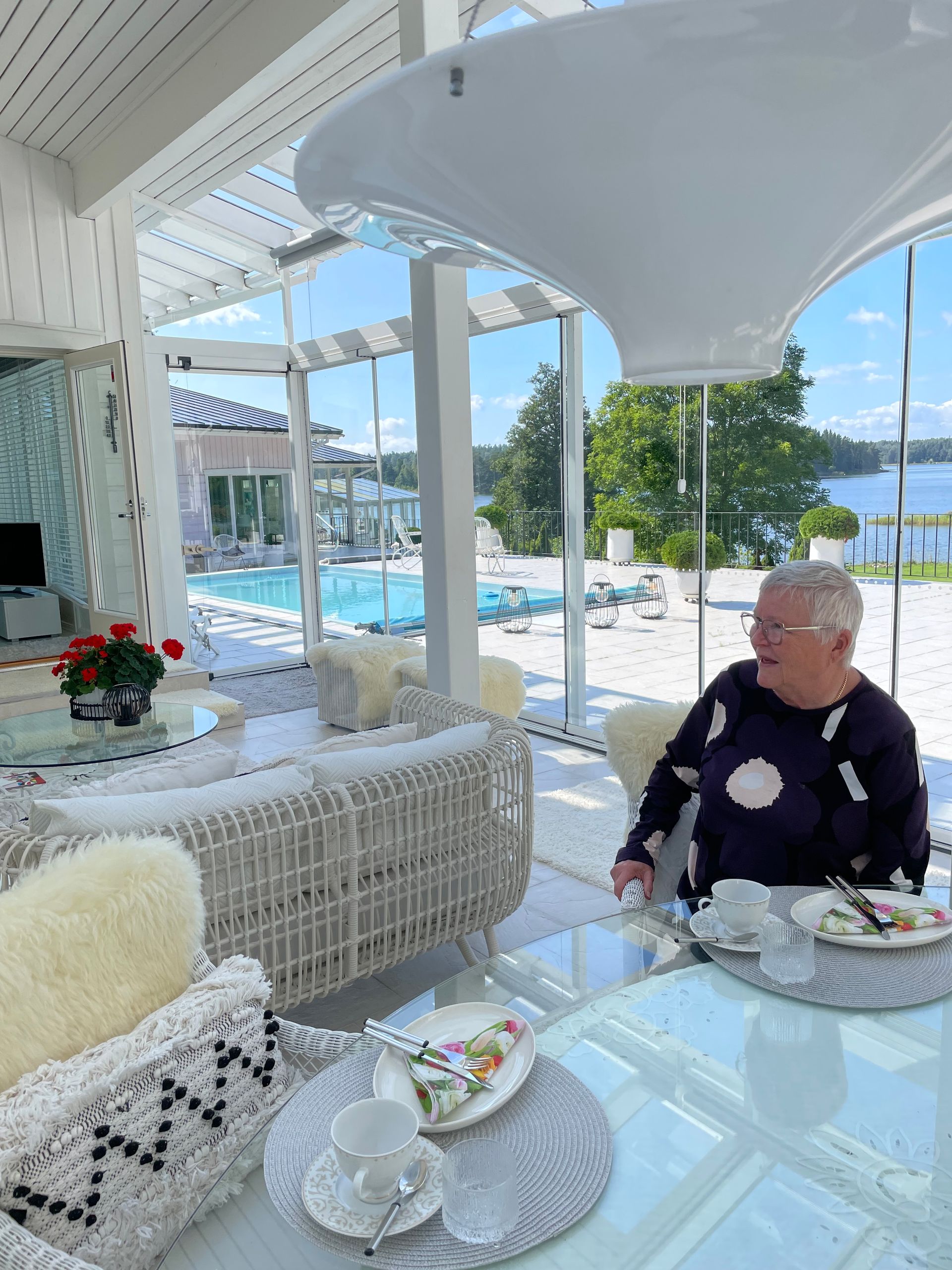 Nainen pöydän ääressä valkoisessa aurinkohuoneessa, uima-allas- ja järvinäköalalla.