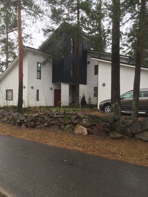 Valkoinen talo mustalla julkisivulla, kivimuuri edessä, puita ja auto.