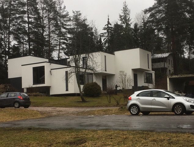 Valkoinen moderni koti, jonka edessä on pysäköitynä autoja, taustalla puita.